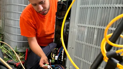 Estes Services Technician in orange shirt repairing and inspecting an outdoor air conditioning unit with wiring and tools.