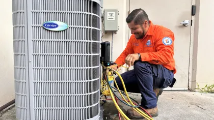 Estes Services Technician in orange uniform repairing a Carrier air conditioning unit with diagnostic tools and colorful cables outdoors.