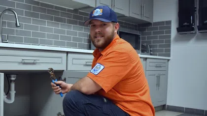 Plumber in orange shirt and cap kneeling by kitchen sink holding a wrench in a tiled kitchen.