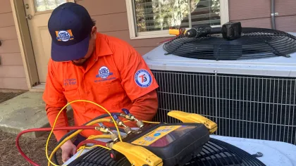 Estes Services Technician in orange uniform servicing an outdoor HVAC unit with diagnostic tools and colored hoses connected.