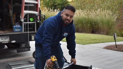 Technician in blue hoodie using a drill to install a metal HVAC unit outdoors near a truck.