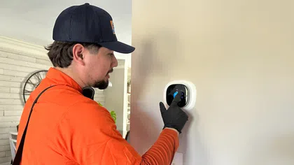 Technician in orange shirt adjusting a smart thermostat on a beige wall inside a home.