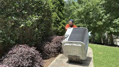 Estes Services Technician in orange shirt and cap moving large HVAC unit on dolly along garden pathway in sunny backyard
