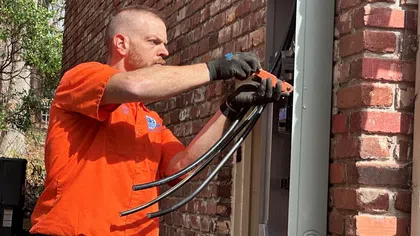 Estes Services Electrician in orange shirt working on electrical panel outdoors next to brick wall during daytime.