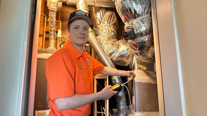 Estes Services Technician in orange shirt inspecting HVAC system pipes and ducts in a utility room with diagnostic tools.