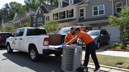 Two technicians unloading a new air conditioning unit from a white truck in a suburban neighborhood on a sunny day