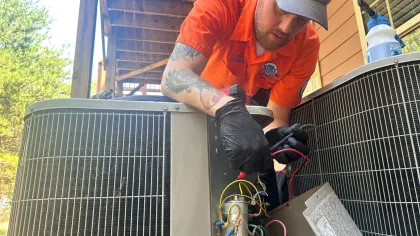 Estes Services AC Repair Technician wearing gloves repairs outdoor air conditioning unit wiring under a porch roof on a sunny day.