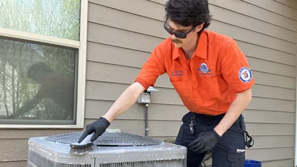 Technician in orange shirt cleaning a Carrier air conditioning unit outside a modern house.
