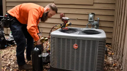 Estes Services Technician in orange shirt servicing a Rheem air conditioning unit outside a beige home with tools on top.