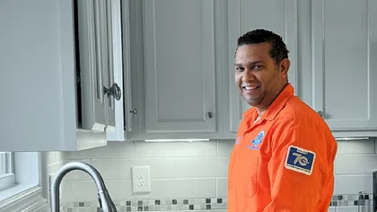Smiling Estes plumber in orange uniform standing by kitchen sink with plumbing tool on granite countertop.