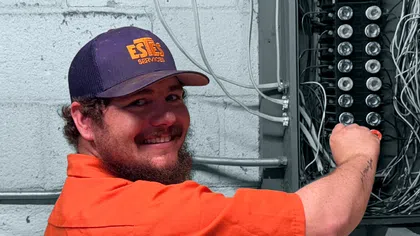 Electrician in orange shirt checking circuit breaker panel with wires in a basement setting.