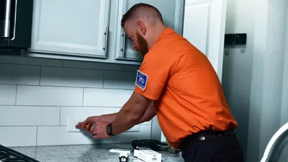 Technician in orange shirt installing or repairing a kitchen electrical outlet near stove and cabinets