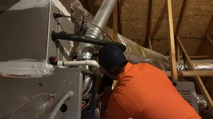 Estes Services Technician in an orange uniform inspecting clogs in a furnace system inside an attic with wooden beams and ductwork.