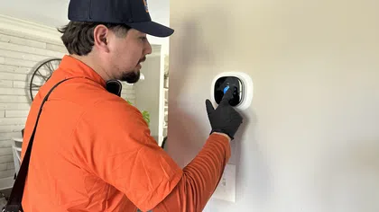 Technician in orange shirt adjusting a smart thermostat on a beige wall inside a home.