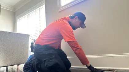 Estes Services Technician in orange shirt and black gloves inspecting floor vent inside a bright modern room.