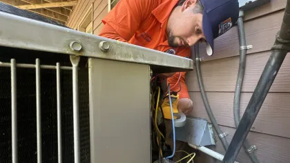 Estes Services Technician in orange shirt and cap inspecting and repairing an air conditioning unit outdoors near house siding.