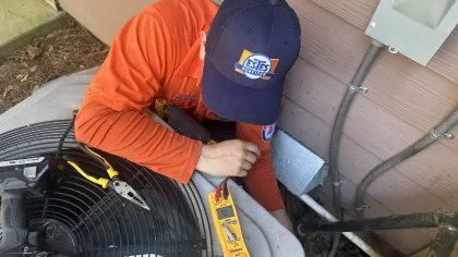 Estes Services HVAC technician in orange shirt and cap using a yellow meter to check an air conditioner unit outside a house.