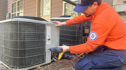Estes Services Technician in orange uniform inspecting a Carrier air conditioning unit and using diagnostic equipment outdoors.
