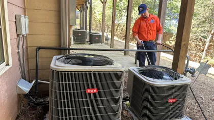 Estes Services Technician in orange shirt servicing Bryant air conditioning units outside a residential building under a covered porch.