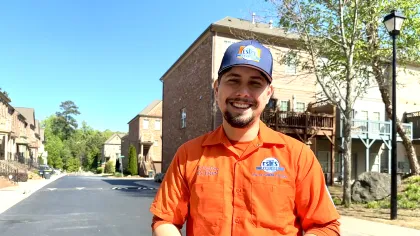 Smiling  Estes Hvac Technician  in orange uniform and cap standing on suburban street holding a smartphone on a sunny day