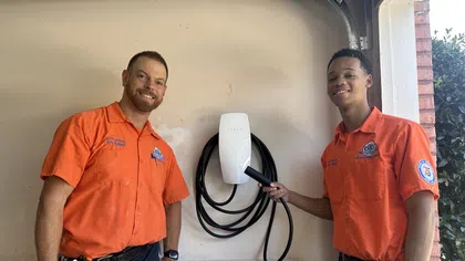 Two Estes Services technicians in orange shirts install a Tesla electric vehicle charger on a beige garage wall.