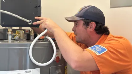 Estes Plumber Technician in orange uniform fixing and inspecting a water heater with tools and pipes in a utility room.