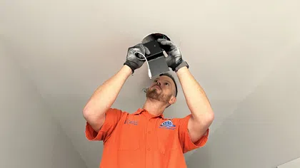 Electrician in orange shirt installing a ceiling light fixture while wearing black gloves inside a room.