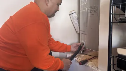 Estes Plumber in orange sweater fixing a water heater with a hose and tool inside a utility room.