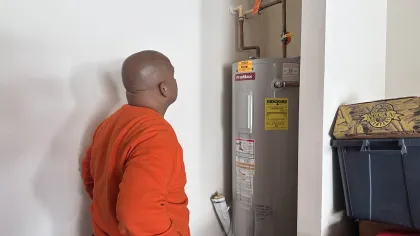 Estes Plumber in orange sweatshirt inspecting a residential ProMax water heater with pipes in a utility room.