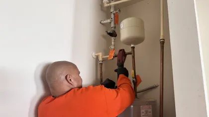 Estes Plumber Technician in orange shirt using a power tool to repair pipes and water heater in a small utility room.
