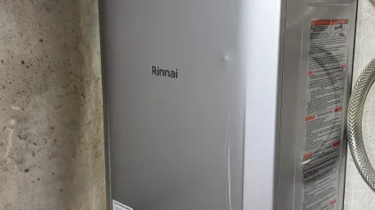 Rinnai tankless water heater mounted on concrete wall with connected pipes and ventilation pipe above.