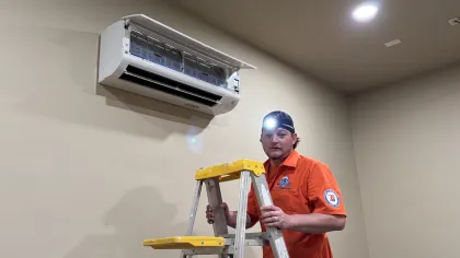 Estes Technician in orange uniform repairing wall-mounted air conditioner while standing on a yellow ladder indoors.