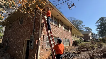 Two Estes Services workers in orange shirts install or repair outdoor cables on a brick house using a tall red ladder on a sunny day.