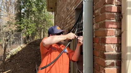 Estes Services Technician in orange shirt and cap connecting cables to an electric panel on a brick wall outdoors.