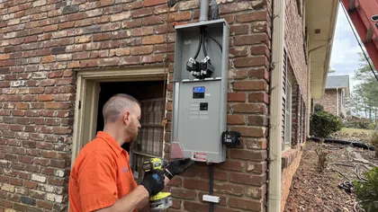 Estes Services Electrician installing or repairing an outdoor electrical panel on a brick house wall with a cordless drill.