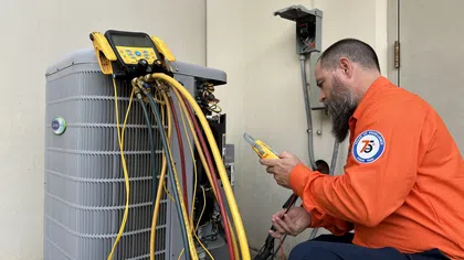 Technician in orange uniform testing HVAC unit with gauges and diagnostic tools beside building wall.