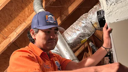 HVAC technician working on furnace system in attic wearing orange uniform and blue cap with ESTES logo.