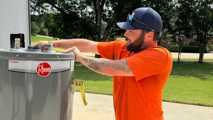 Estes Plumber in orange shirt installing Rheem water heater outdoors on a sunny day near green trees.