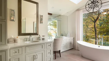 Bright modern bathroom with freestanding tub next to large window and vanity with mirror and chair.