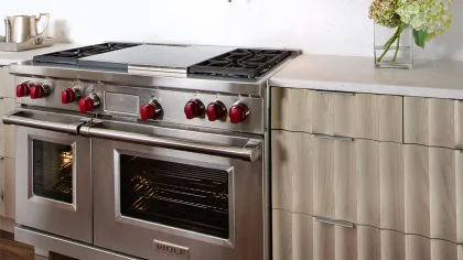 Stainless steel Wolf gas range with red knobs in a modern kitchen with light wood cabinetry and a flower vase.