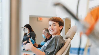 Smiling teenage boy with braces giving a thumbs-up while sitting in a dental chair at a clinic.