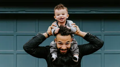 Smiling toddler sitting on father's shoulders in front of blue garage door, both enjoying playful moment.