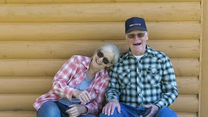 Elderly couple in plaid shirts smiling and laughing, sitting against a wooden cabin wall outdoors.