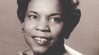 Black and white portrait of a smiling woman with short curly hair wearing a patterned dress and earrings.