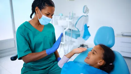 Dentist in scrubs and mask giving a high-five to a smiling child patient in dental chair.
