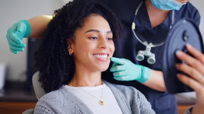 Woman smiling and checking her teeth in a mirror during a dental checkup with a dentist wearing gloves.