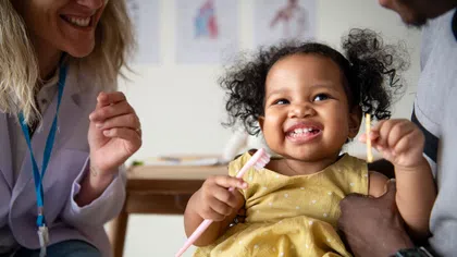 Smiling toddler holding a toothbrush and dental tool with adults in a bright dental clinic.