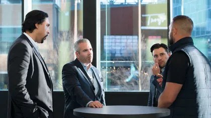 Four men in business attire engaged in a serious discussion around a high table in a modern office space.