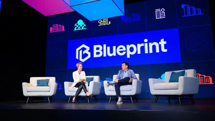 Two people seated on stage armchairs under BluePrint logo with colorful cubes above and building icons on backdrop