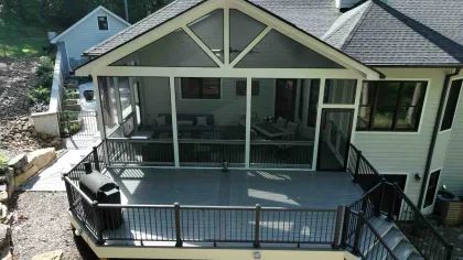Elevated deck with screened porch attached to a white house overlooking a wooded backyard area.